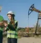 Two engineers using a tablet in front of an oil pump jack, discussing average gas well royalties per acre and using a royalty calculator.
